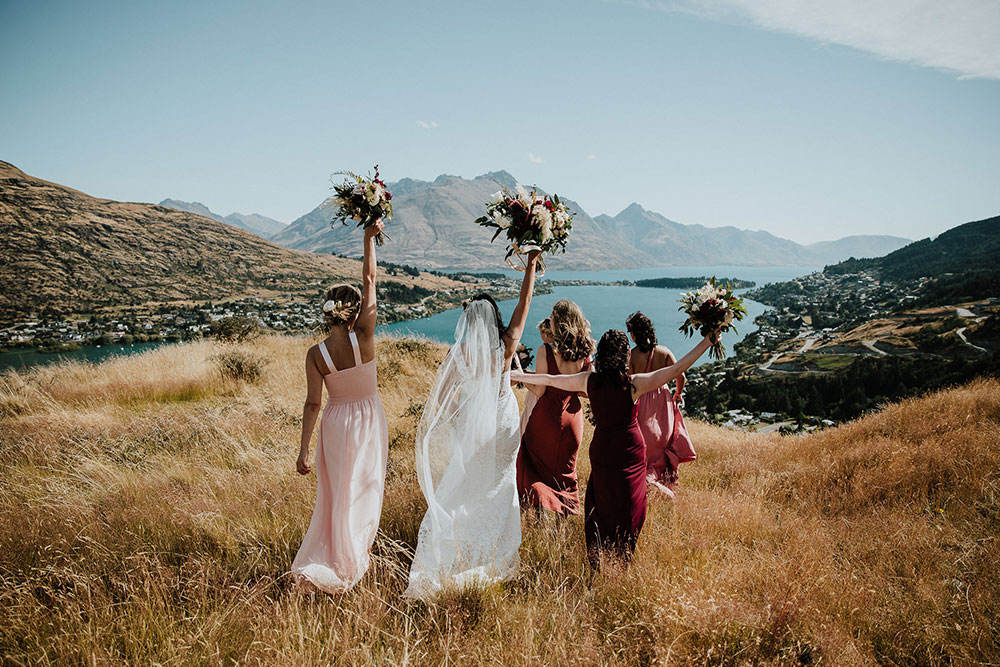Bride and bridesmaids raise flower bouquets in the air overlooking Queenstown. Tipi wedding by Gather and Gold