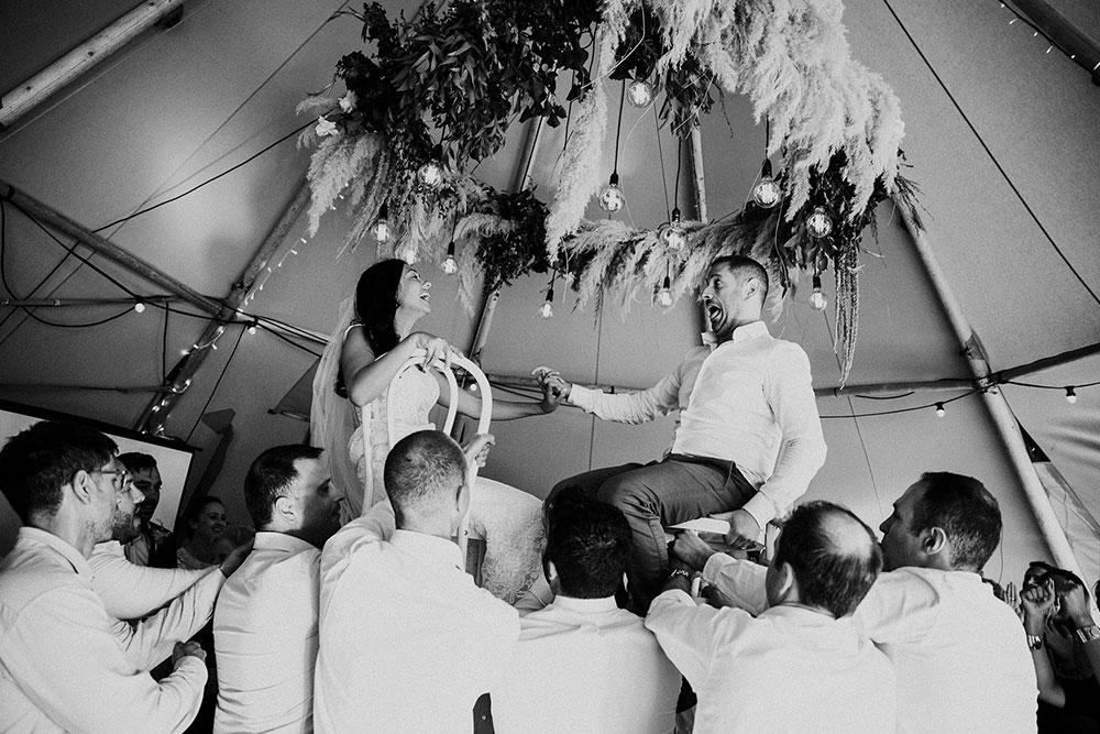 Black and white of bride and groom being raised on chairs by guests inside tipi at wedding - by Gather and Gold