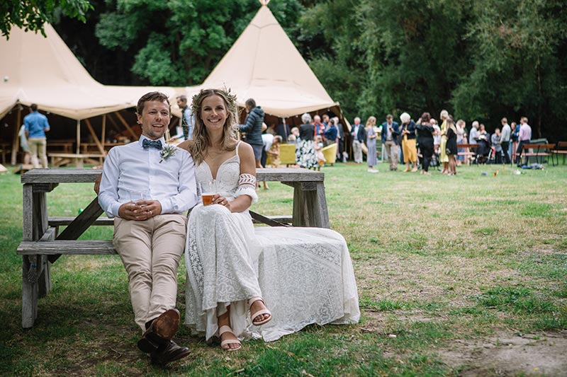 Bride and Groom pose on bench in front of multiple tipi set up for Wanaka tipi wedding by Gather and Gold