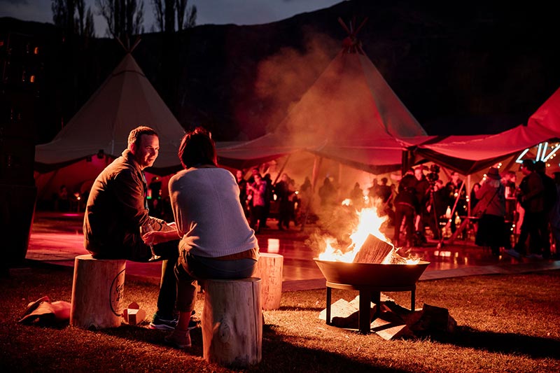 Two people sit by a fire pit at the NZME corporate event with Gather and Gold tipis