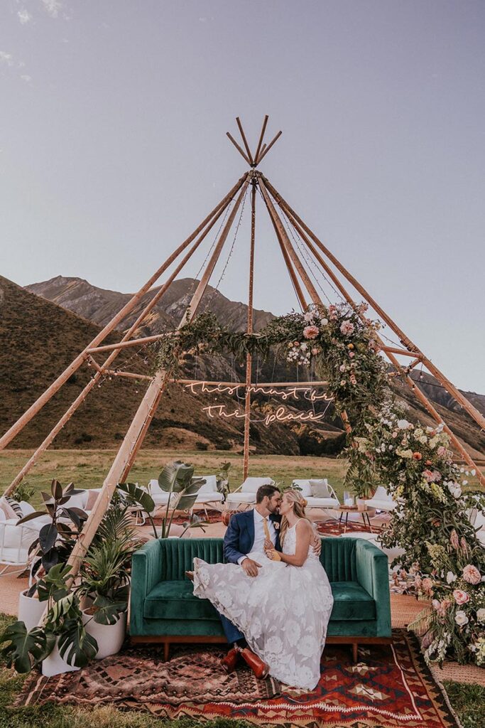 Wooden frame tipi decorated with dried flowers and seating underneath. Bride and groom embrace