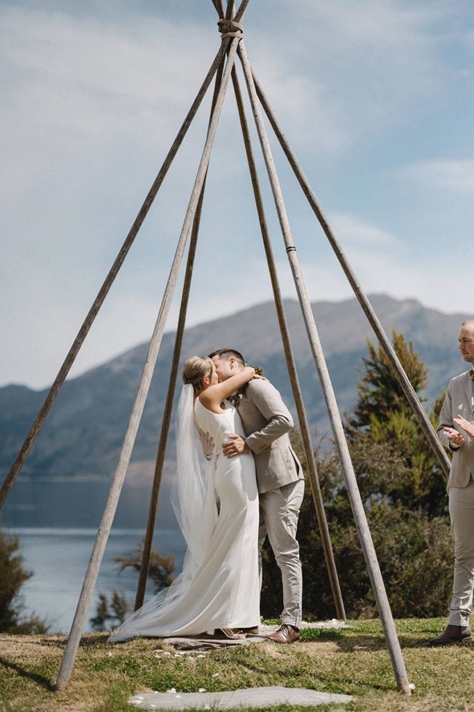 Fairytale tipi wedding in Hawea, South Island, New Zealand. Bride and groom kiss under bare tipi frame with lake backdrop.