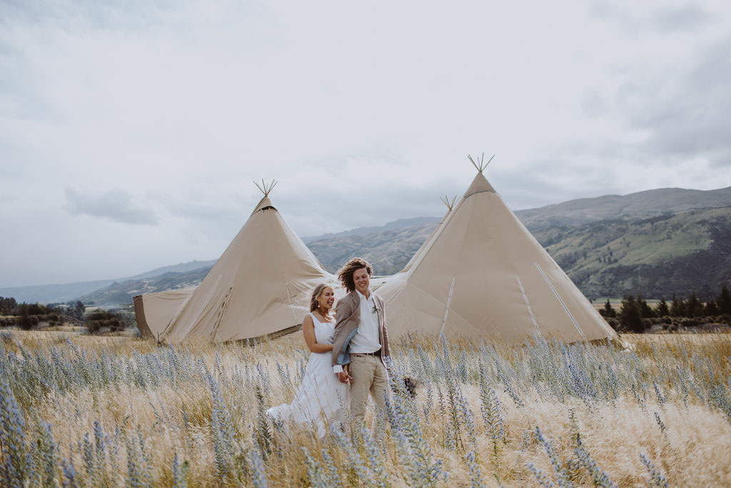 Bride and groom walk through south island field of flowers with 2 large tipis in the background - top tips for a seamless wedding day from Gather & Gold tipi hire