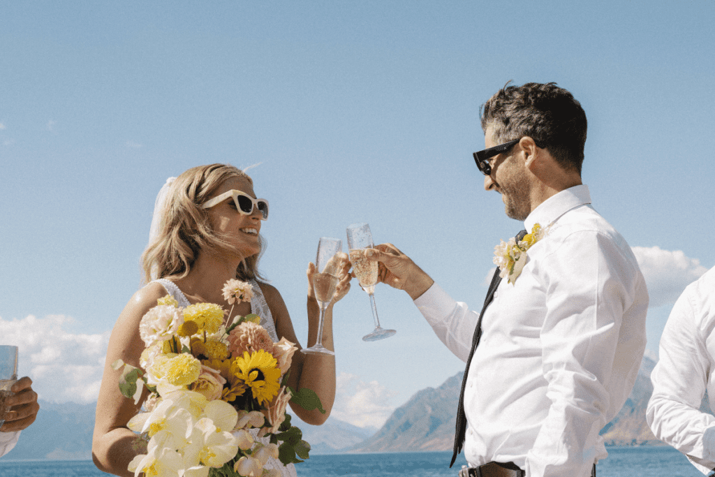 A couple in wedding attire clink champagne glasses under a blue sky