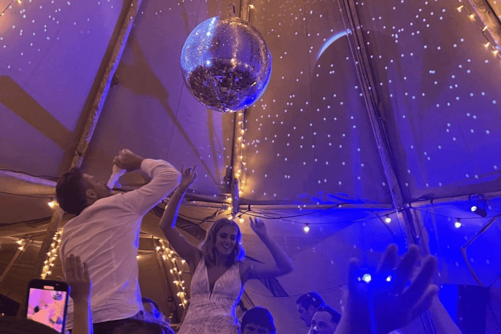 A lady with blonde hair and in a wedding dress dances under a disco ball in a Tipi