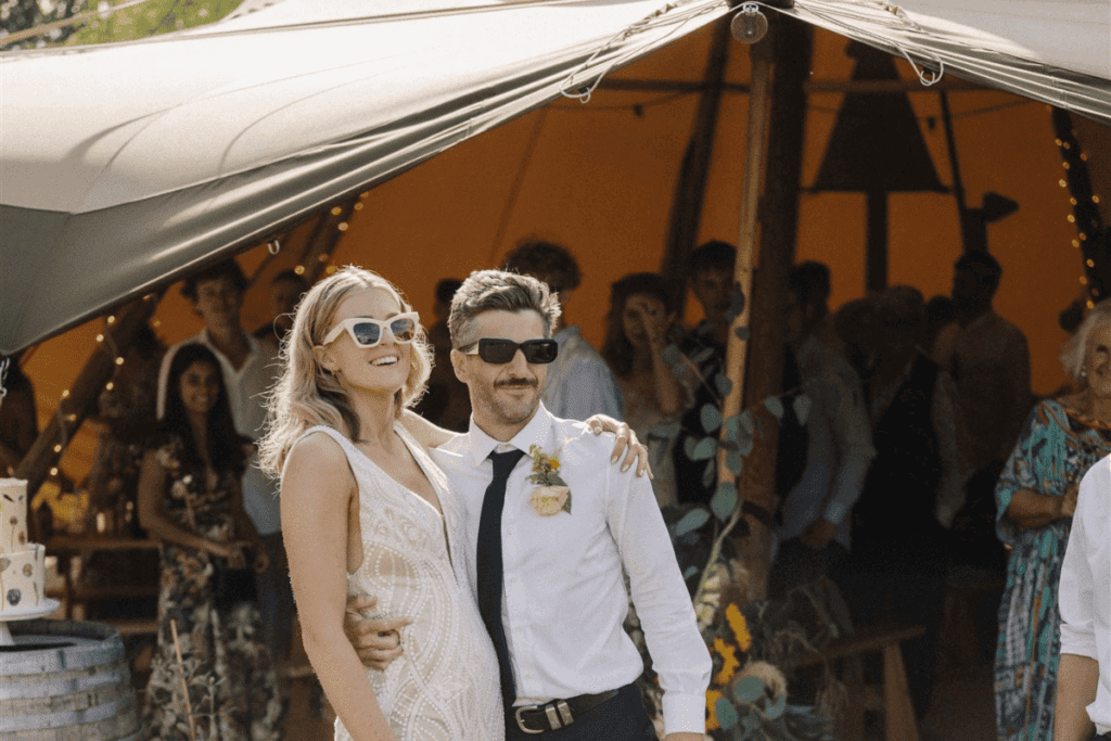 A couple in wedding attire stand in front of a Tipi with sunglasses on