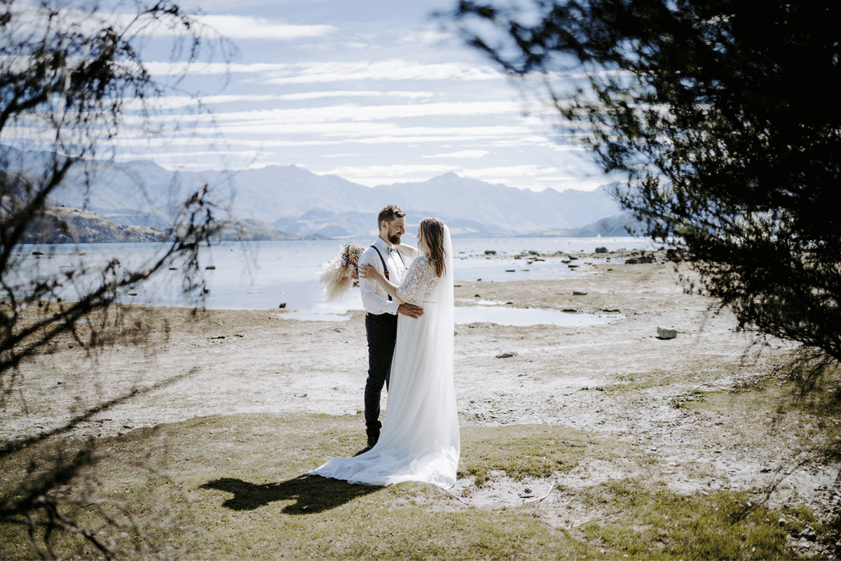A bride and groom pose in front of a lake and mountains in Wanaka
