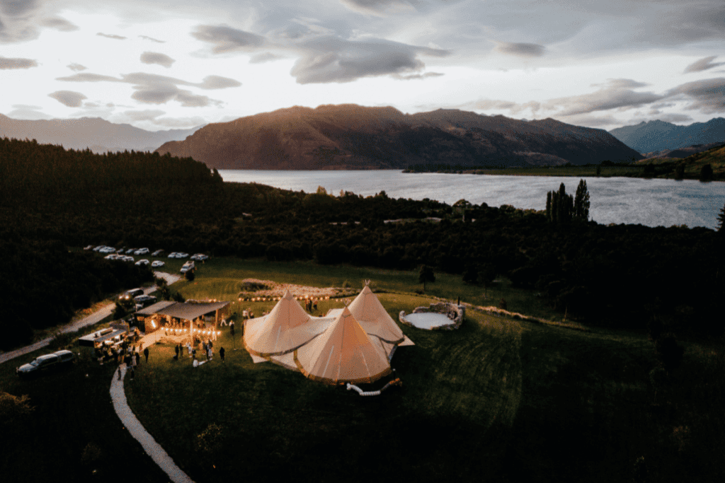Three Nordic tipis sit in a field next to a lake at dusk in New Zealand