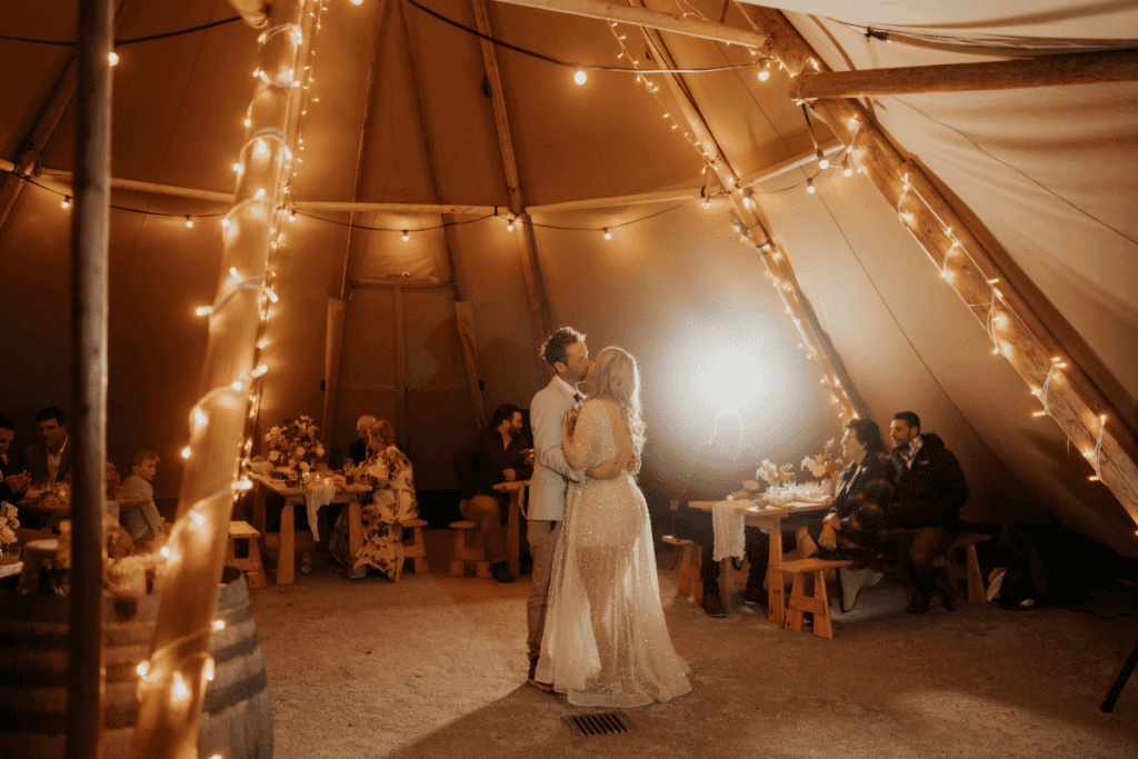 A bride and groom share a moment of intimacy on the dance floor, illuminated by strands of fairy lights in a cozy tipi, with guests seated at tables around them enjoying the festive atmosphere.