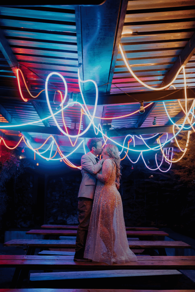 A couple sharing a kiss stands on a picnic table under a canopy of colorful neon lights, creating an enchanting and romantic atmosphere.