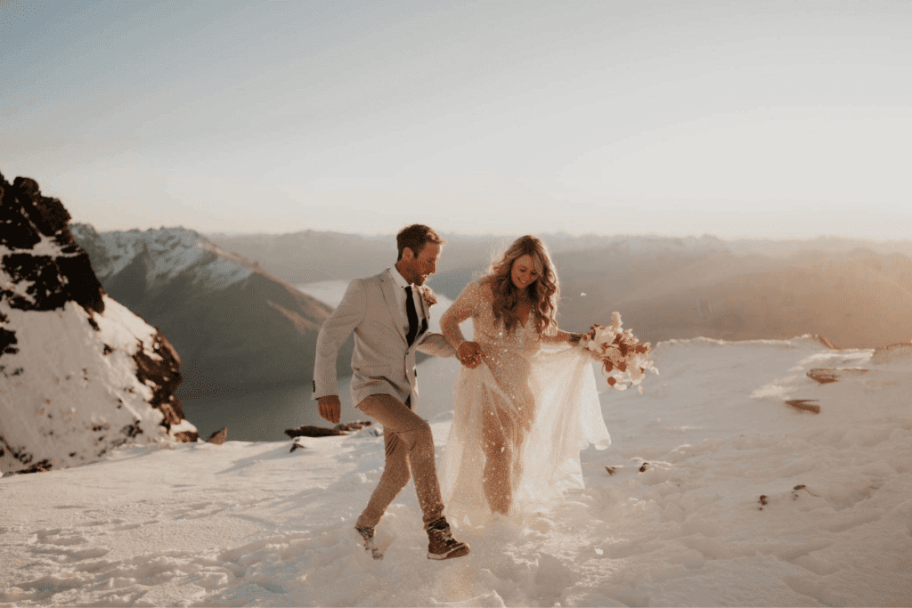 A bride in a sparkling gown holding a bouquet and a groom in a white suit gaze towards the setting sun on a snowy mountain, with a tranquil lake and mountain range in the background.