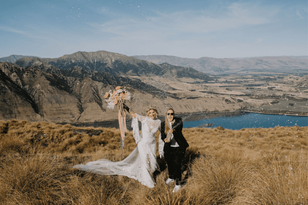 Two joyful brides celebrate by popping a champagne bottle on a scenic mountaintop in Wanaka, with sweeping views of a valley, lake, and distant mountains under a clear blue sky. One bride holds a bouquet of flowers, and both are dressed in wedding attire, radiating happiness.