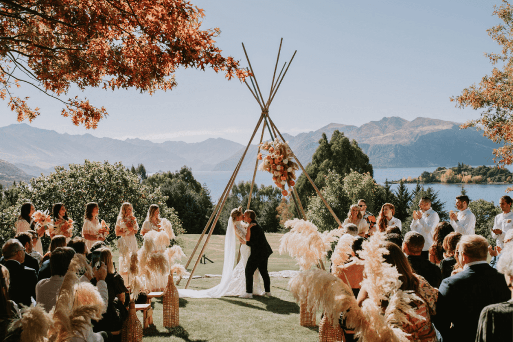 A beautiful outdoor wedding ceremony with two brides sharing a kiss under a tipi-style arbor adorned with flowers. The setting features lush greenery, pampas grass decorations, and a stunning backdrop of mountains and a lake. Guests, including bridesmaids and groomsmen, are seated and standing, witnessing the joyous moment on a clear, sunny day.