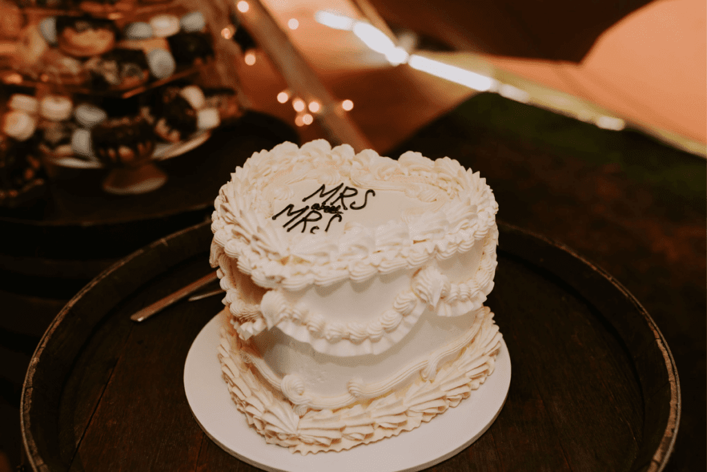 A white wedding cake with intricate icing details and the text "Mrs and Mrs" written on top. The cake is placed on a wooden barrel, with a dessert table featuring various treats visible in the background, softly illuminated by string lights.