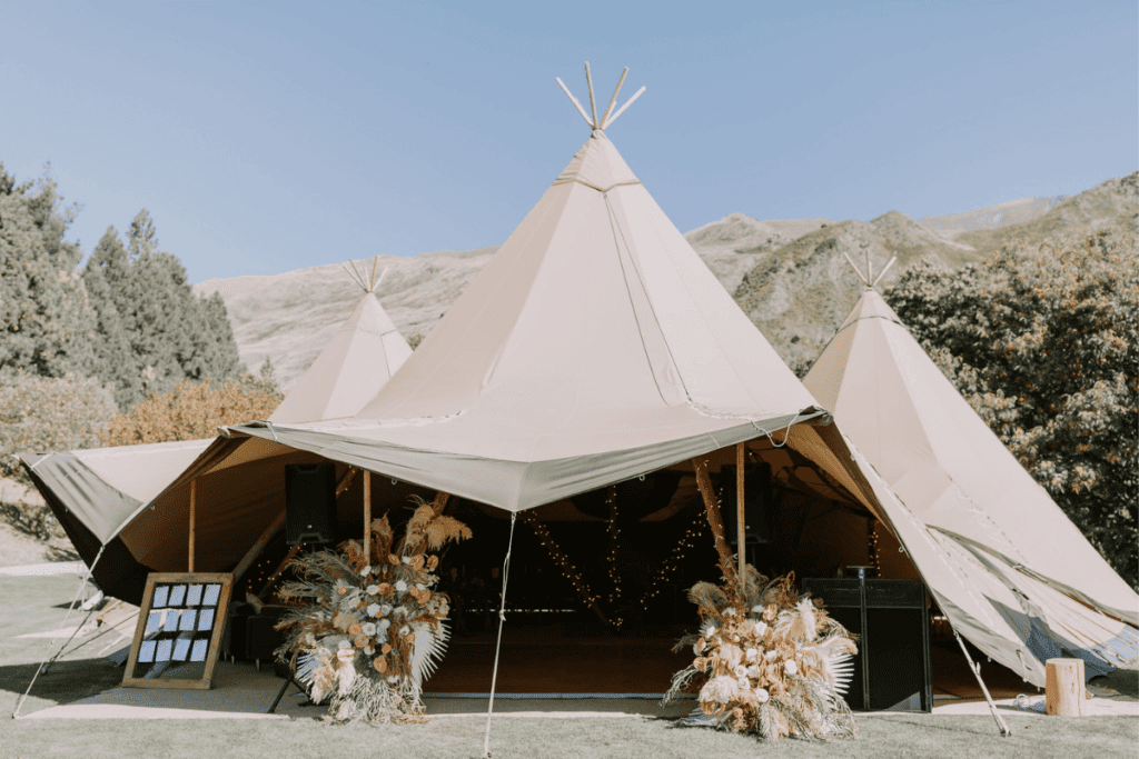 Three large Nordic tipis set up outdoors against a backdrop of mountains and trees, forming a spacious and elegant wedding venue in Wanaka. The entrance is decorated with floral arrangements featuring pampas grass and other dried flowers, and there is a seating chart displayed on an easel. The tipis are open, revealing warm, inviting lighting inside.