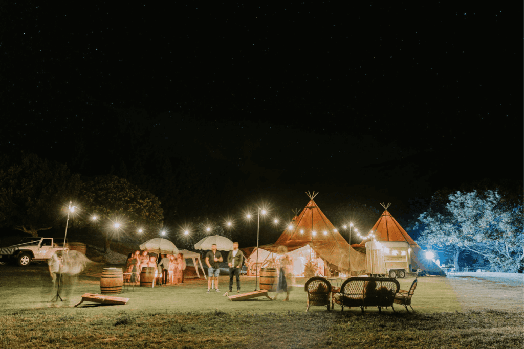 Three illuminated Nordic tipis set against a starry night sky, forming a cozy and enchanting wedding reception area. String lights and festoon lighting create a warm atmosphere, with guests mingling and enjoying the outdoor setting. Various seating arrangements, including wicker furniture and barrels, add to the rustic charm.